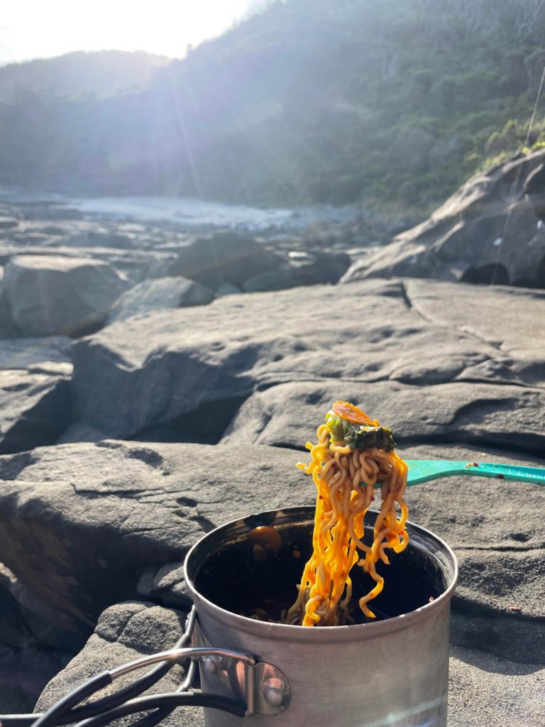 Noodles held on a spoon out of a hiking pot with the rocks in the background and coastline in the distance. The sun is starting to set.