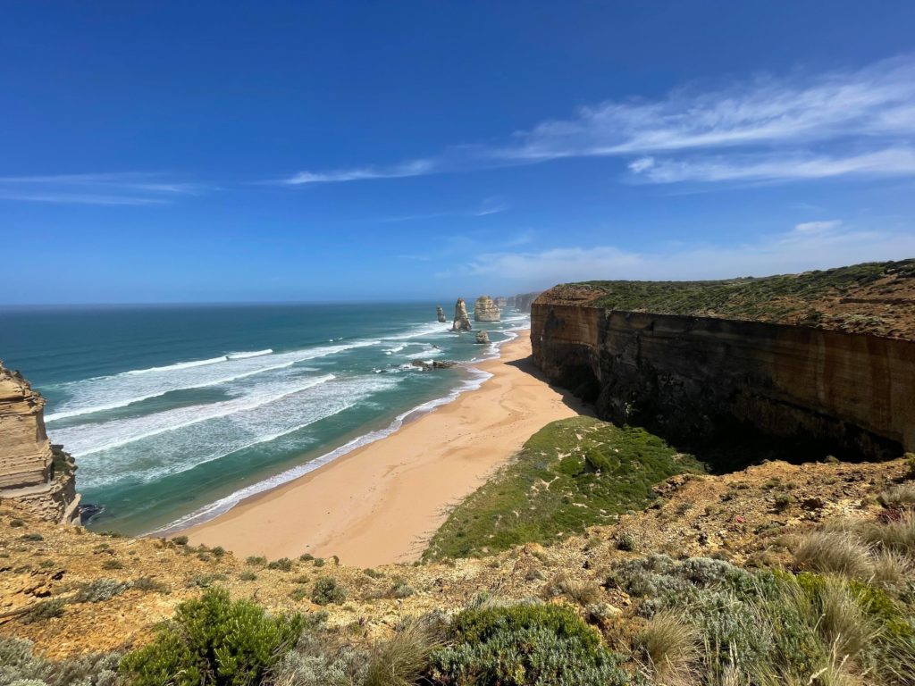 The 12 Apostles are in the background. In the foreground is a grassy cliff overlooking the beach and ocean. There is a large cliff following alongside the beach on the right side of the picture. The sky is blue with a few wispy clouds.