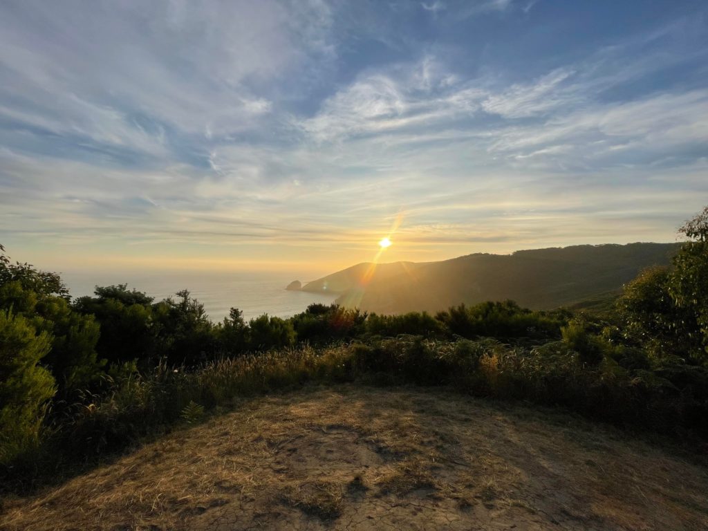 A grassy cleared hill with a stunning view of the ocean sky and sun starting to set over escarpments.