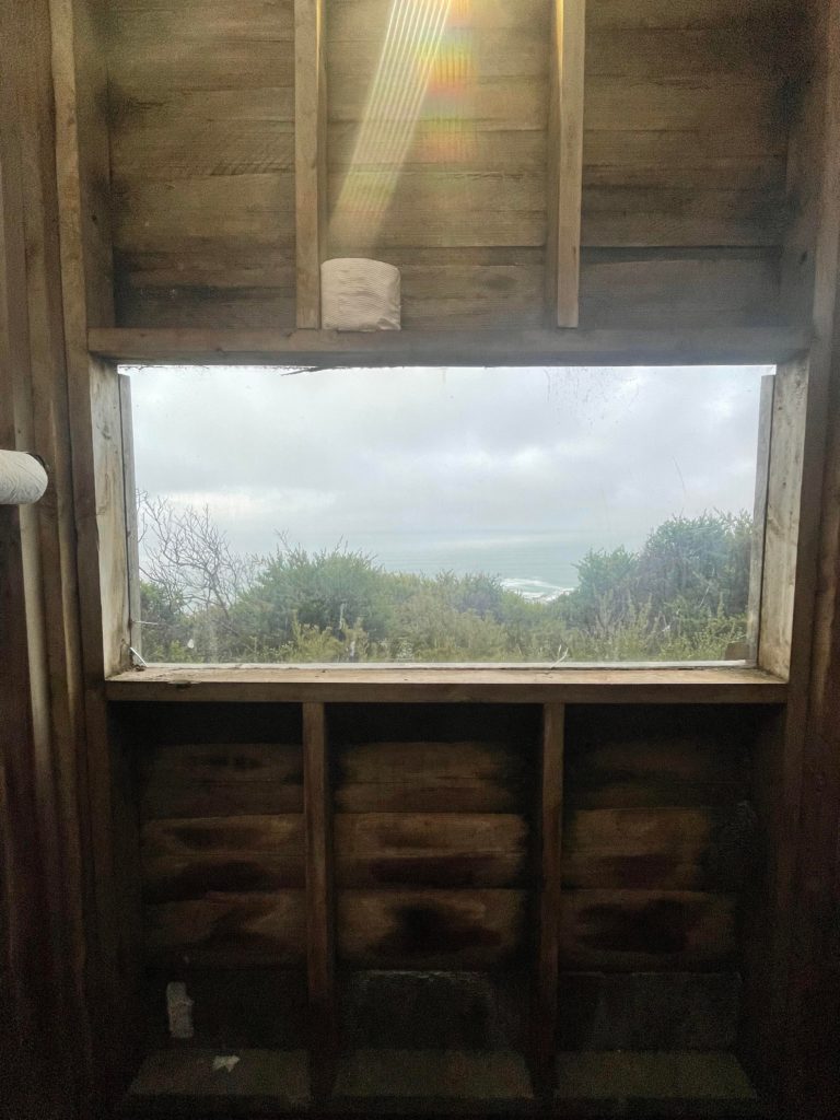 An image of the window out of a drop toilet looking at the spectacular vegetation and ocean.