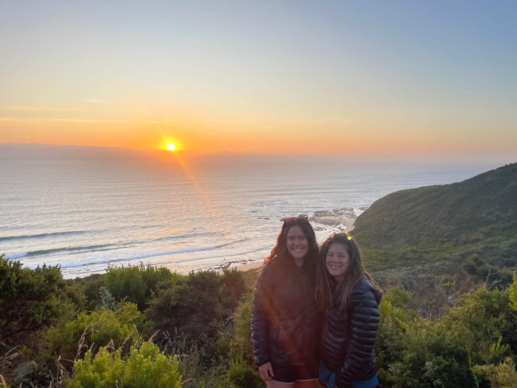 Two women embracing for a photo in front of the ocean sunset.