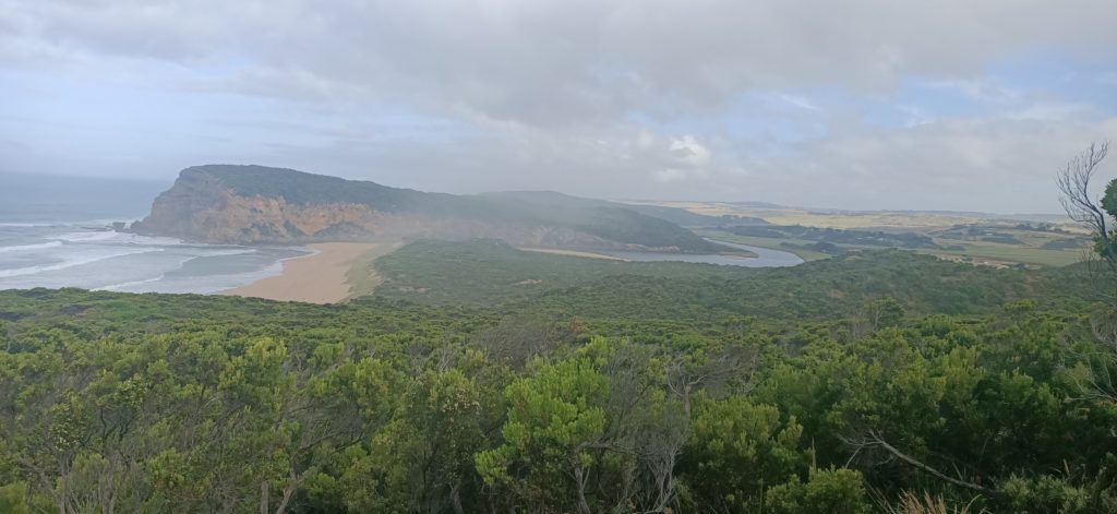 The mouth of the coastal Gellibrand River. The sky is hazy and cloudy. The coastal bush is green and lush, there is an escarpment in the back ground and you can also start to see the edge of princetown.