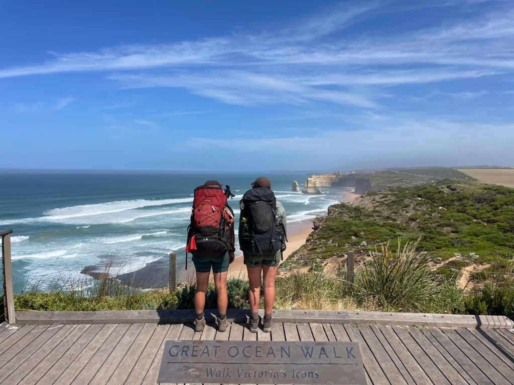 A lookout with a plack for the Great Ocean Walk. Two women with their backs to the camera, carrying heavy packs stand in front of it. The background is of the 12 apostles and the ocean. The sky is blue with wispy clouds.