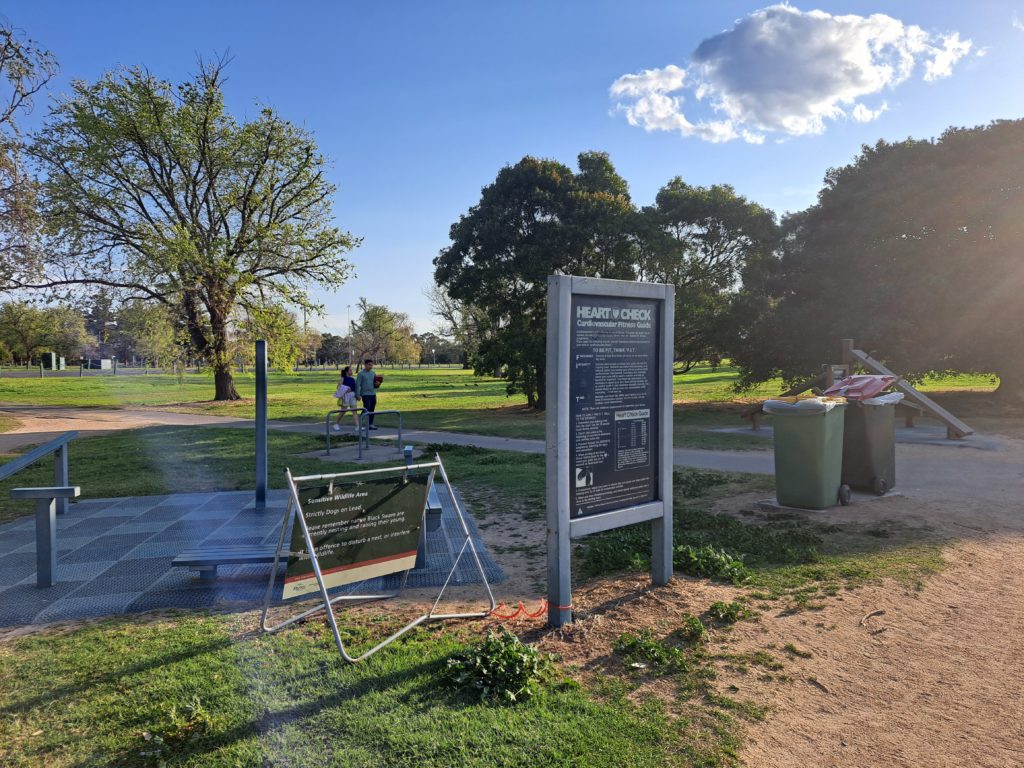 there is a sign describing how to use the workout equipment that is behind it. In the background a trees and people walking passed on a concrete path.
