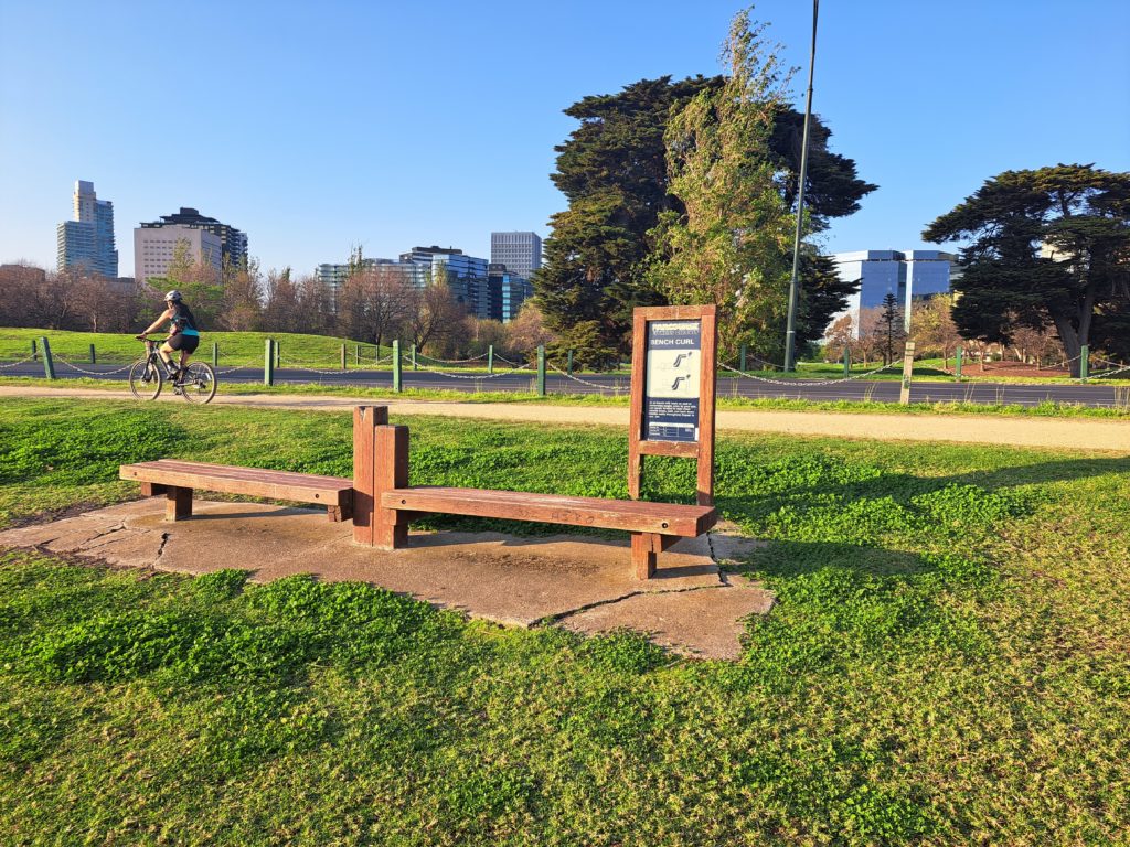 There are wooden benches for a workout zone surrounded by grass with an instruction plaque behind it. There is a road and then the city skyline in the background.