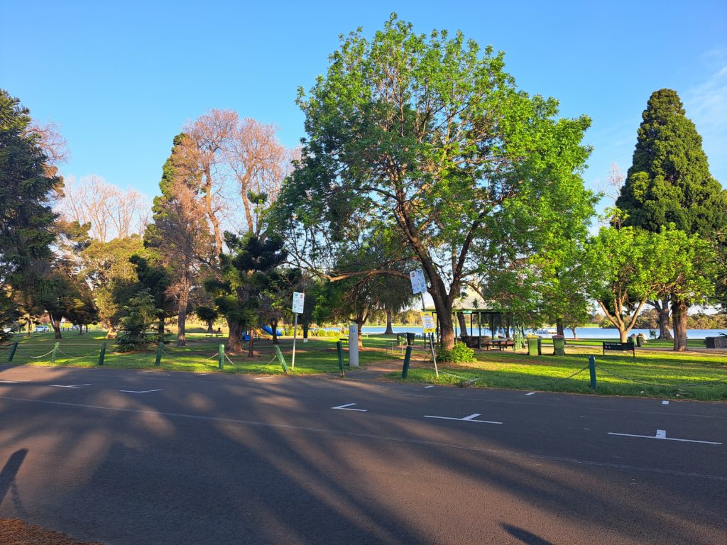 The carpark is in the foreground with no cars inside. There are a variety of trees behind the carpark and the lake is in the distance. 