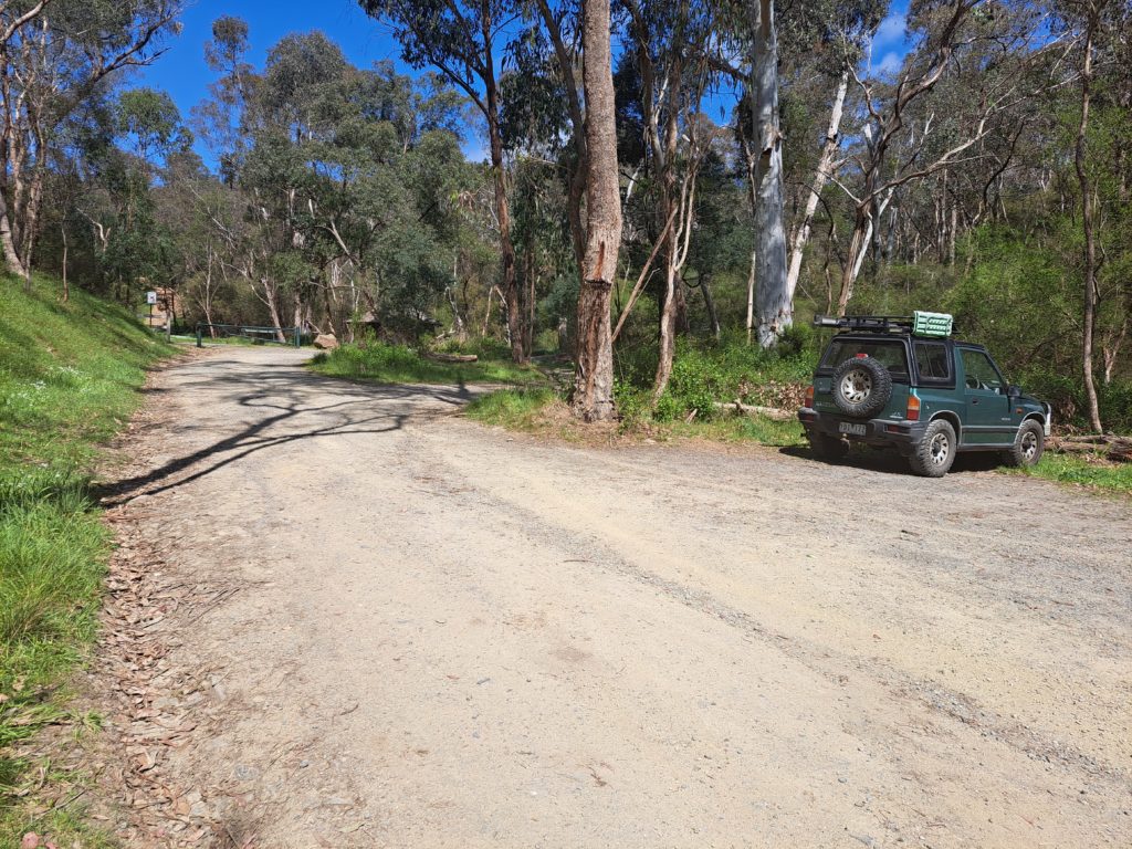 View of gravel road and carpark before the turning cycle of Whipstick Gully Road. There is a green suzuki vitara parked in one spot to the right of the photo.