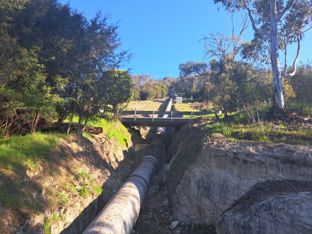 A view from the bridge below the Lower Rubicon Power Station pipeline. There is the road crossing the pipeline and then a grassy hill.